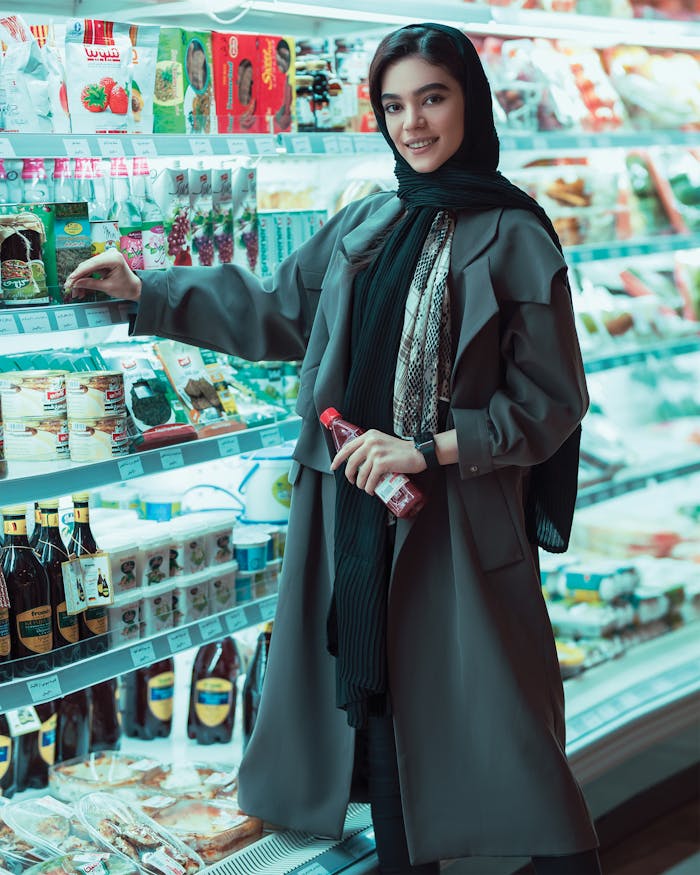 A stylish woman in a headscarf shopping in a well-lit supermarket aisle, smiling as she examines products.