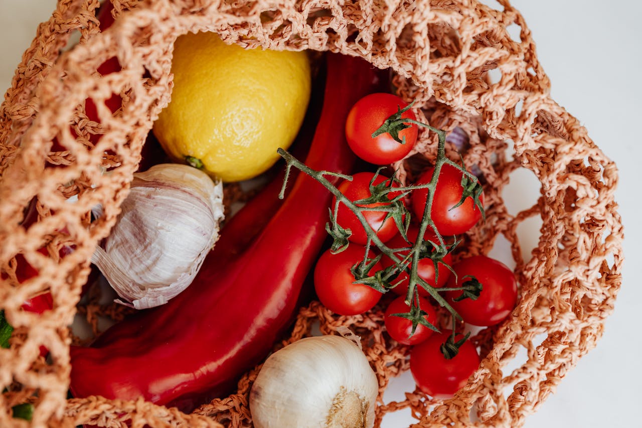 Top view of fresh produce in a reusable bag, featuring tomatoes, garlic, lemon, and peppers, promoting healthy eating.