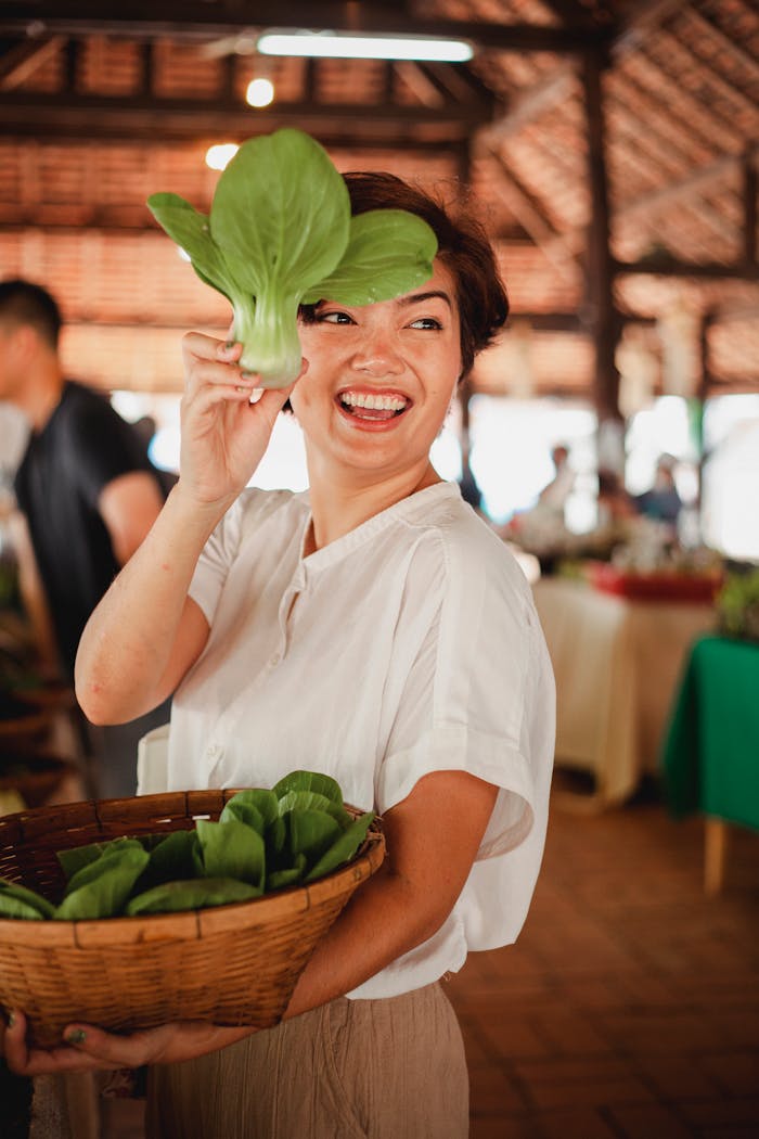 Smiling woman with fresh greens and basket at a local farmers market, enjoying organic produce.